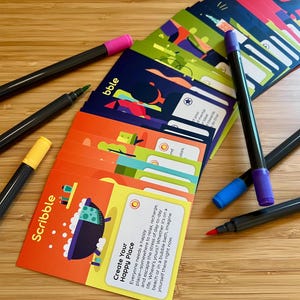 May include: A collection of colorful cards with the word "Scribble" and "Create Your Happy Place" printed on them. Several black markers with colored tips, including pink, green, yellow, blue, and purple, are scattered around the cards. The cards have various illustrations.