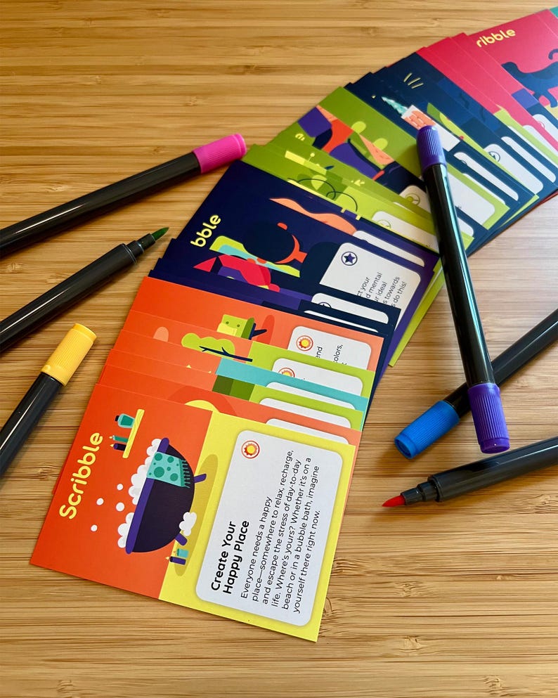 May include: A collection of colorful cards with the word "Scribble" and "Create Your Happy Place" printed on them. Several black markers with colored tips, including pink, green, yellow, blue, and purple, are scattered around the cards. The cards have various illustrations.