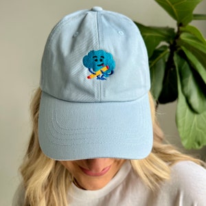 Może przedstawiać: Jasno błękitna czapka z daszkiem z haftowanym rysunkiem chmury trzymającej ołówek. Chmura ma niebieską twarz z żółtym uśmiechem i czerwonymi oczami.