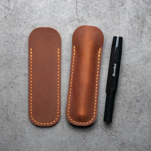 Puede incluir: Dos fundas para bolígrafos de cuero marrón con costuras amarillas, y un bolígrafo negro con la palabra "Lamy" impresa en él.