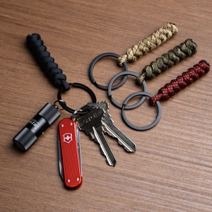 Puede incluir: Un conjunto de llaveros con anillos de metal negro y asas de paracord trenzado en negro, beige, verde y rojo. Incluye una pequeña linterna negra, una navaja suiza roja y llaves.