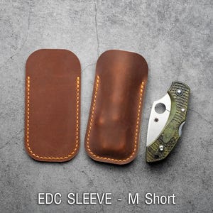 May include: Two brown leather sleeves for carrying a pocket knife. The sleeves are stitched with a light brown thread. The text "EDC SLEEVE - M Short" is printed below the sleeves.