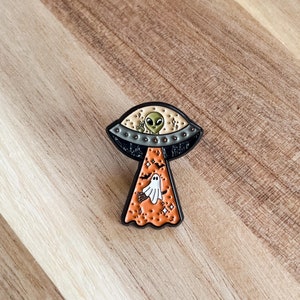 May include: A Halloween-themed enamel pin featuring a UFO with an alien giving a peace sign. The UFO emits an orange beam with a ghost and bats. The pin is black, orange, white, and green.