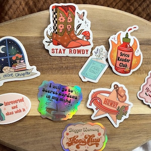 K&ouml;nnte beinhalten: Ein Set aus neun bunten Aufklebern mit verschiedenen Designs und Texten. Die Aufkleber beinhalten ein Paar Cowboystiefel, ein Buch mit dem Text "Fairy Smut", eine scharfe Sauce mit dem Text "Spicy Reader Club", ein Buch mit dem Text "I will go to Secret Gardens in my mind", ein Buch mit dem Text "One more chapter", einen Cowboyhut mit dem Text "Howdy", ein Raumschiff mit dem Text "Introverted and okay with it", ein Buch mit dem Text "Trigger Warning Book Club" und ein Buch mit dem Text "Proud Spicy Reader".