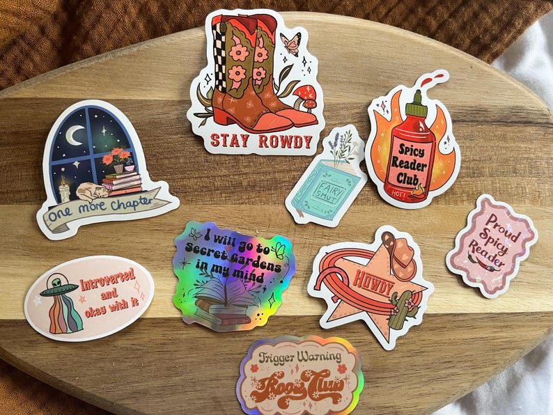 K&ouml;nnte beinhalten: Ein Set aus neun bunten Aufklebern mit verschiedenen Designs und Texten. Die Aufkleber beinhalten ein Paar Cowboystiefel, ein Buch mit dem Text "Fairy Smut", eine scharfe Sauce mit dem Text "Spicy Reader Club", ein Buch mit dem Text "I will go to Secret Gardens in my mind", ein Buch mit dem Text "One more chapter", einen Cowboyhut mit dem Text "Howdy", ein Raumschiff mit dem Text "Introverted and okay with it", ein Buch mit dem Text "Trigger Warning Book Club" und ein Buch mit dem Text "Proud Spicy Reader".