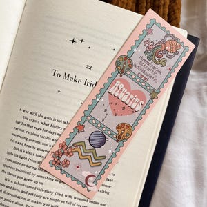 May include: A pink bookmark with an Aquarius design, showing planets, stars, and flowers. The bookmark has the words "Aquarius" and "HUMANITARIAN, ECCENTRIC, INDIVIDUALISTIC, COOL" printed on it. The bookmark is in an open book.