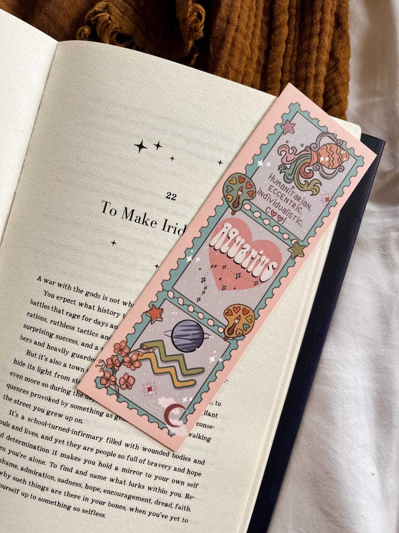 May include: A pink bookmark with an Aquarius design, showing planets, stars, and flowers. The bookmark has the words "Aquarius" and "HUMANITARIAN, ECCENTRIC, INDIVIDUALISTIC, COOL" printed on it. The bookmark is in an open book.