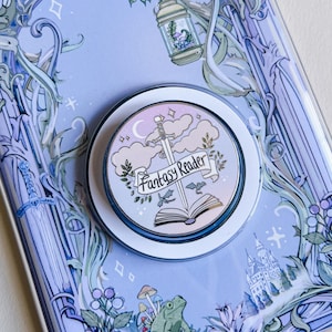 Op de afbeelding: Een ronde, wit en zilverkleurige popsocket met een fantasiethema. Het ontwerp toont een zwaard, wolken, een banner met "Fantasy Reader" en een open boek. De achtergrond is lichtblauw met bloemen- en kasteelillustraties.