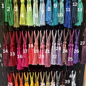 May include: A display of 40 decorative tassels in various colours, each with a hanging loop. The tassels are arranged in rows, displaying a wide range of hues, including greens, blues, reds, pinks, yellows, and browns. The image is well-lit, showing the texture and detail of the tassels.