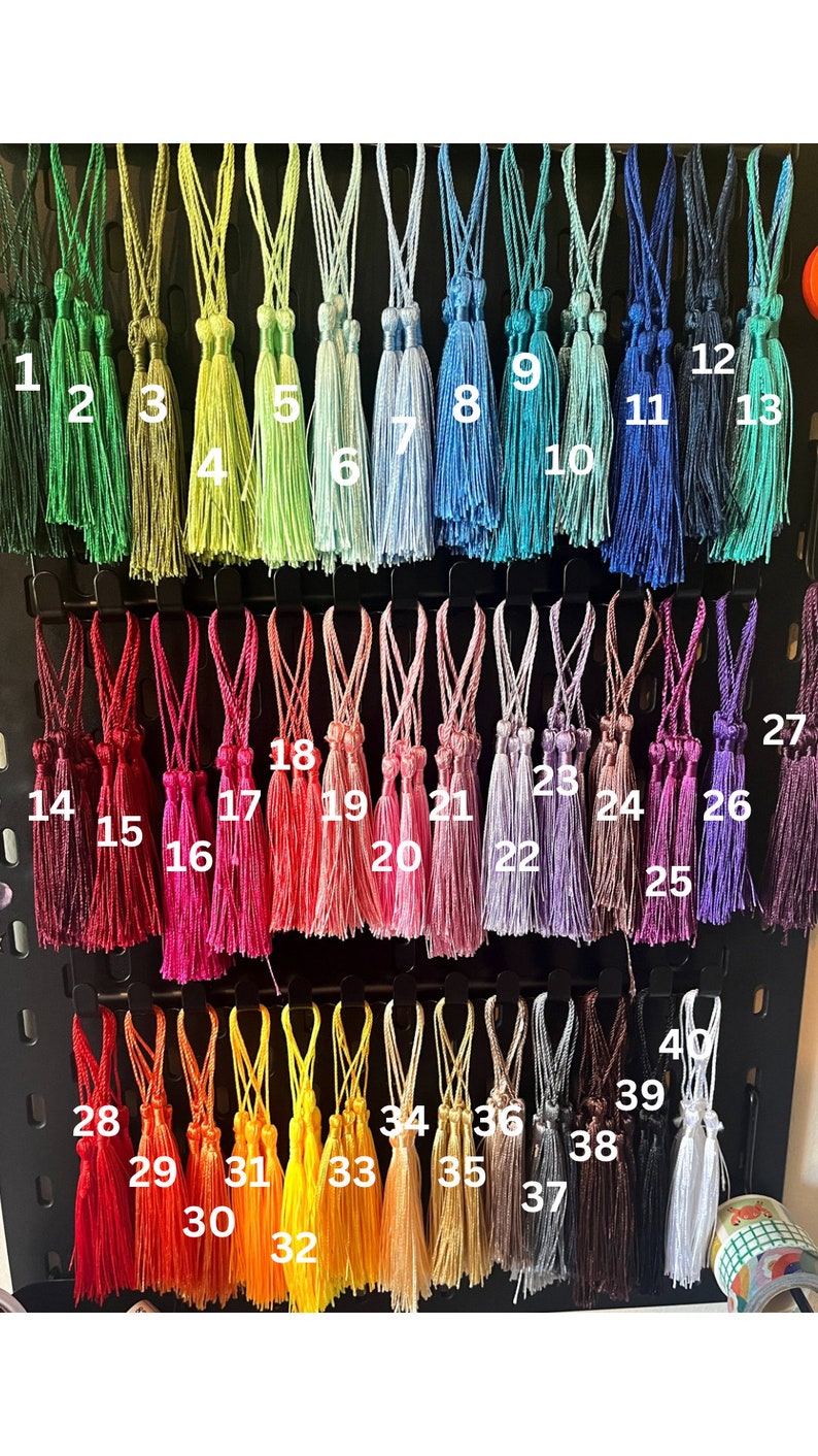May include: A display of 40 decorative tassels in various colours, each with a hanging loop. The tassels are arranged in rows, displaying a wide range of hues, including greens, blues, reds, pinks, yellows, and browns. The image is well-lit, showing the texture and detail of the tassels.