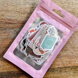 K&ouml;nnte beinhalten: Eine rosa wiederverschlie&szlig;bare Plastikt&uuml;te mit einem transparenten Fenster, das eine Vielzahl von Aufklebern zeigt. Die Aufkleber zeigen Illustrationen von B&uuml;chern, Blumen, Sternen und dem Text "Fairy Smut".