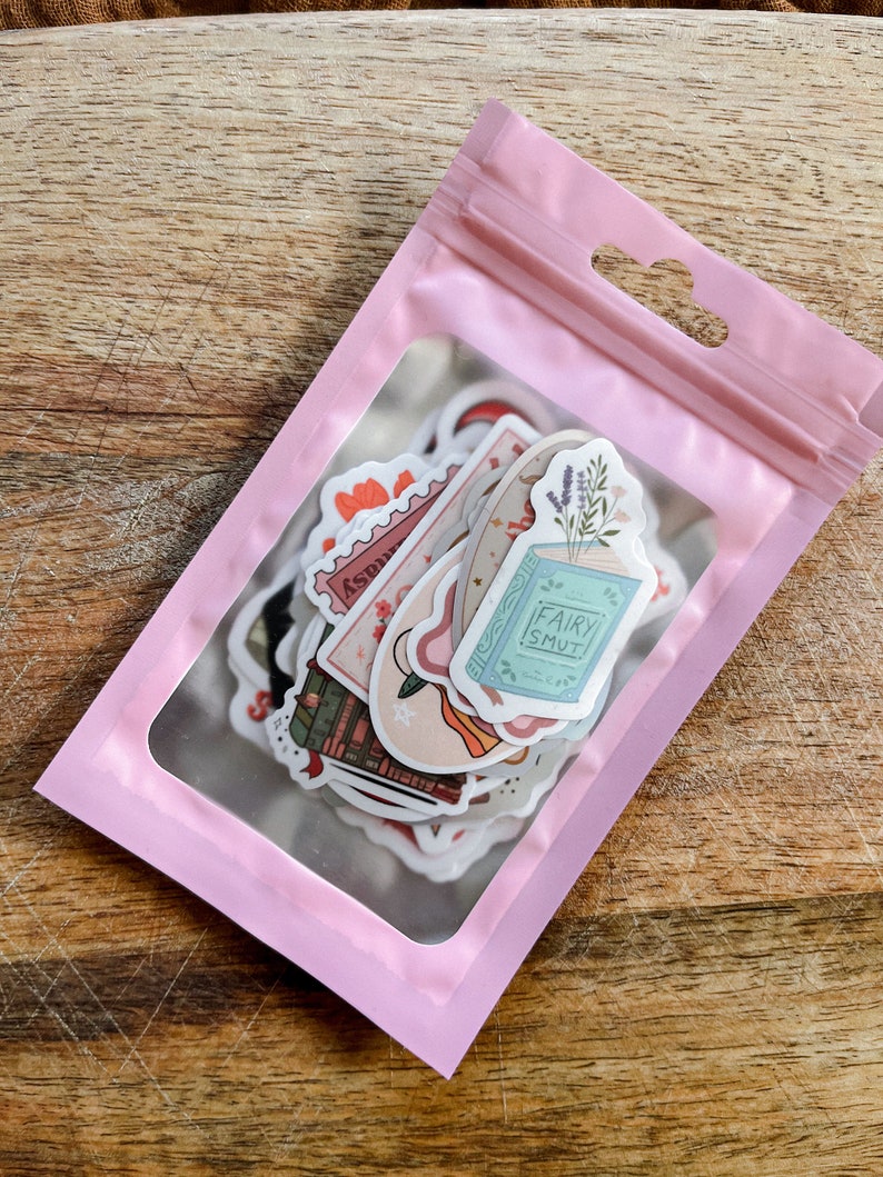 K&ouml;nnte beinhalten: Eine rosa wiederverschlie&szlig;bare Plastikt&uuml;te mit einem transparenten Fenster, das eine Vielzahl von Aufklebern zeigt. Die Aufkleber zeigen Illustrationen von B&uuml;chern, Blumen, Sternen und dem Text "Fairy Smut".