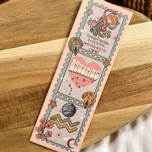 May include: A pink bookmark with the Aquarius zodiac sign. The design includes illustrations of a fish, a planet, and the Aquarius symbol. Text includes "Humanitarian, Eccentric, Individualistic, c&hearts;♡" and "Aquarius".