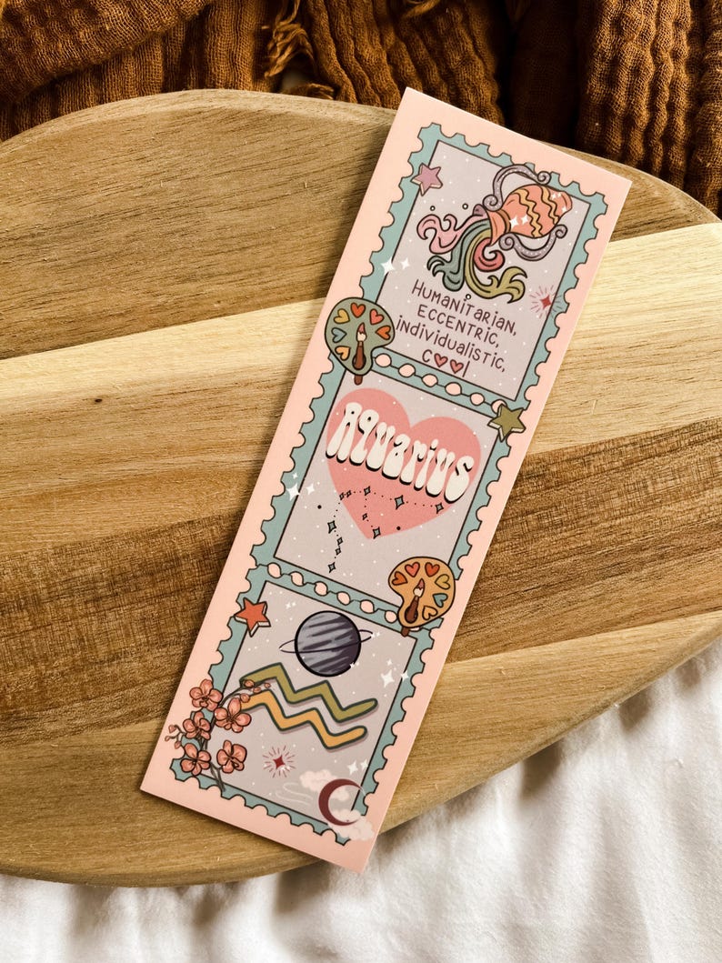 May include: A pink bookmark with the Aquarius zodiac sign. The design includes illustrations of a fish, a planet, and the Aquarius symbol. Text includes "Humanitarian, Eccentric, Individualistic, c&hearts;♡" and "Aquarius".