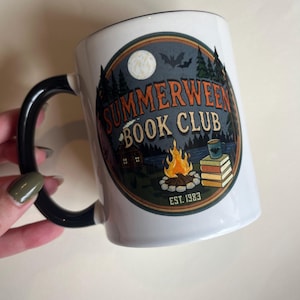 Op de afbeelding: Een witte keramische mok met een zwart handvat en binnenkant. De voorzijde bevat een cirkelvormige afbeelding met een volle maan, vleermuizen, een bos, een hut, een kampvuur en een stapel boeken. De tekst luidt "SUMMERWEEN BOOK CLUB" en "EST. 1983".