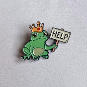 Op de afbeelding: Een groene kikker-pin met een gouden kroon met een rood hart. De kikker houdt een bord vast met de tekst "HELP" in zwarte letters. De pin heeft een glanzende afwerking en staat tegen een witte achtergrond.