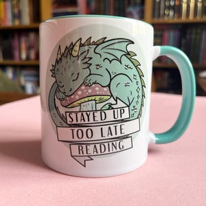 Pode incluir: Caneca de cerâmica branca com alça e borda turquesa. A caneca apresenta uma ilustração de um dragão turquesa adormecido em um cogumelo rosa e branco. O texto na caneca diz "STAYED UP TOO LATE READING."