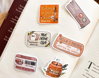 May include: A collection of stickers featuring various designs, including a book club sticker with the text "Enemies to lovers club", a true crime club sticker with a ghost, a sticker that says "Hello I prefer men written by women", a sticker that says "TBR calling cat denied", and a sticker that says "Book Smell" with a candle and floral design.