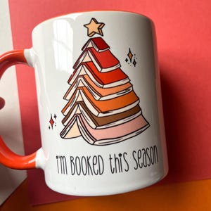 May include: White ceramic mug with a red rim and handle. The mug features a colorful illustration of a Christmas tree made of stacked books with a star on top. The text "I'm booked this season" is printed below the tree.
