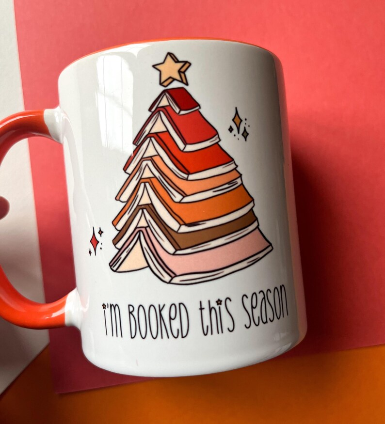 May include: White ceramic mug with a red rim and handle. The mug features a colorful illustration of a Christmas tree made of stacked books with a star on top. The text "I'm booked this season" is printed below the tree.