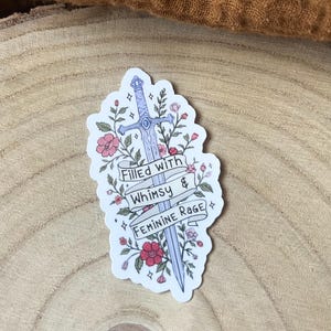 May include: A die-cut sticker featuring a light blue sword entwined with pink and red flowers and green leaves. A white banner wraps around the sword with the black text "Filled with Whimsy & FEMININE RAGE" printed in three distinct lines.