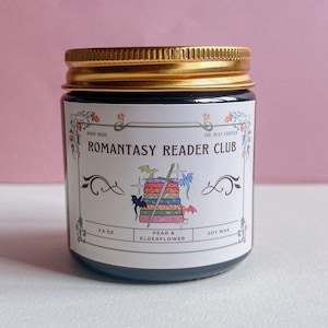 Pode incluir: Um frasco de vidro com tampa dourada, rotulado "ROMANTASY READER CLUB". O rótulo apresenta uma pilha de livros com uma espada e dragões. O frasco contém 99g de cera de soja e é perfumado com pera e flor de sabugueiro.