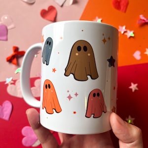 Op de afbeelding: Witte keramische mok met een kleurrijk spookpatroon. De spoken zijn bruin, oranje, roze en blauw. De mok heeft een witte handgreep.