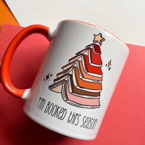 May include: White ceramic mug with an orange handle. The mug features a graphic of a Christmas tree made of stacked books with a star on top. The text "I'm booked this season" is printed on the mug.