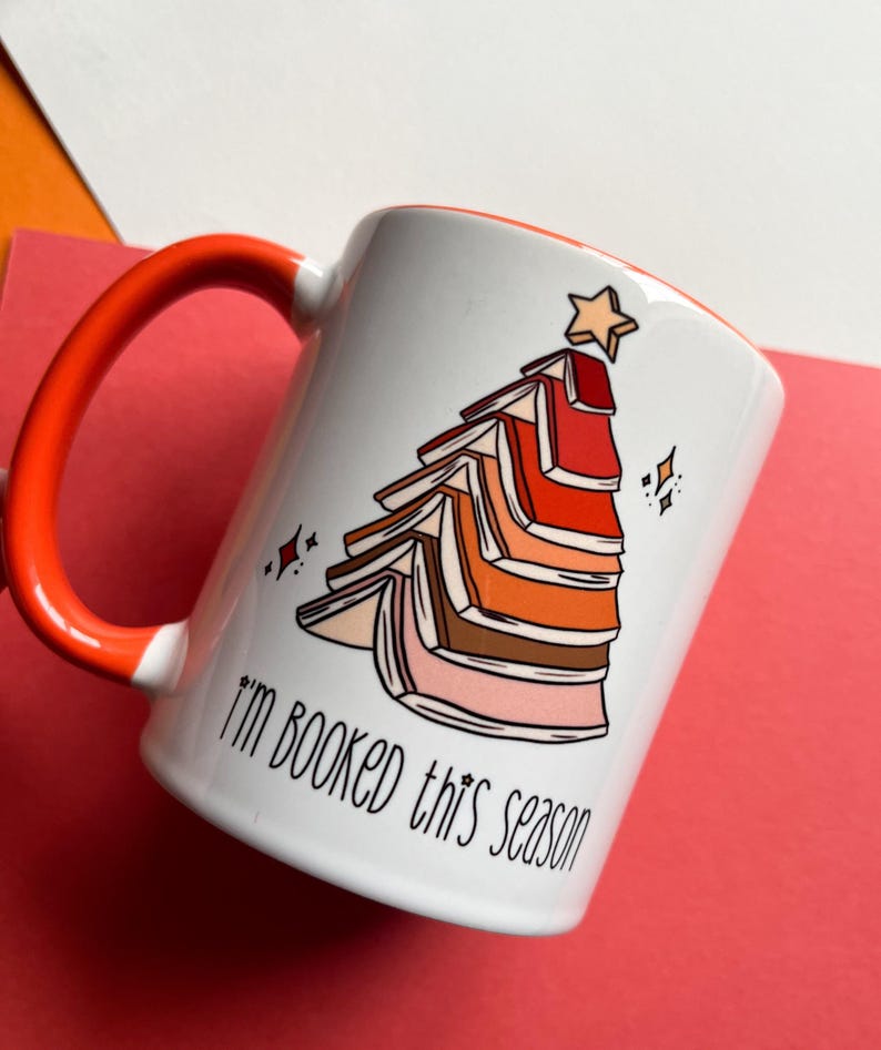 May include: White ceramic mug with an orange handle. The mug features a graphic of a Christmas tree made of stacked books with a star on top. The text "I'm booked this season" is printed on the mug.