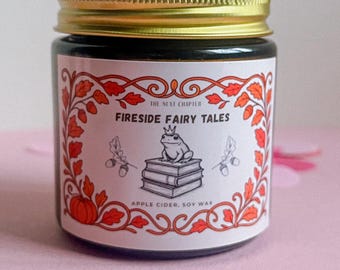 May include: A glass jar candle with a gold lid. The label reads "Fireside Fairy Tales" with a frog on books illustration. The label has an orange and red floral design and the text "Apple Cider, Soy Wax".