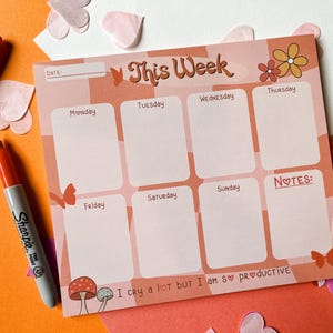 May include: A pink and orange weekly planner with a floral design and the text "This Week". The planner has spaces for each day of the week, Monday through Sunday, and a notes section. The planner also has the text "I cry a lot but I am so productive" at the bottom.