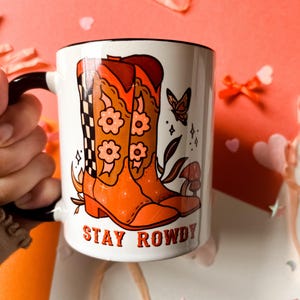 May include: White ceramic mug with a black handle. The mug features a graphic of two orange cowboy boots with floral designs and a butterfly. The text "STAY ROWDY" is printed in red below the boots.