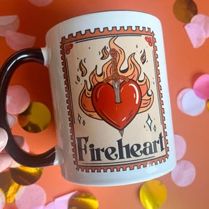 May include: White ceramic mug with a black handle. The mug features a graphic of a red heart with a sword through it, engulfed in flames. The text "Fireheart" is printed below the graphic.
