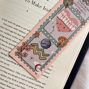 May include: A bookmark with an Aquarius design. The bookmark has a pink border and a light blue interior. The design includes a heart with the word "Aquarius", stars, planets, and other astrological symbols. The bookmark is placed on top of an open book.