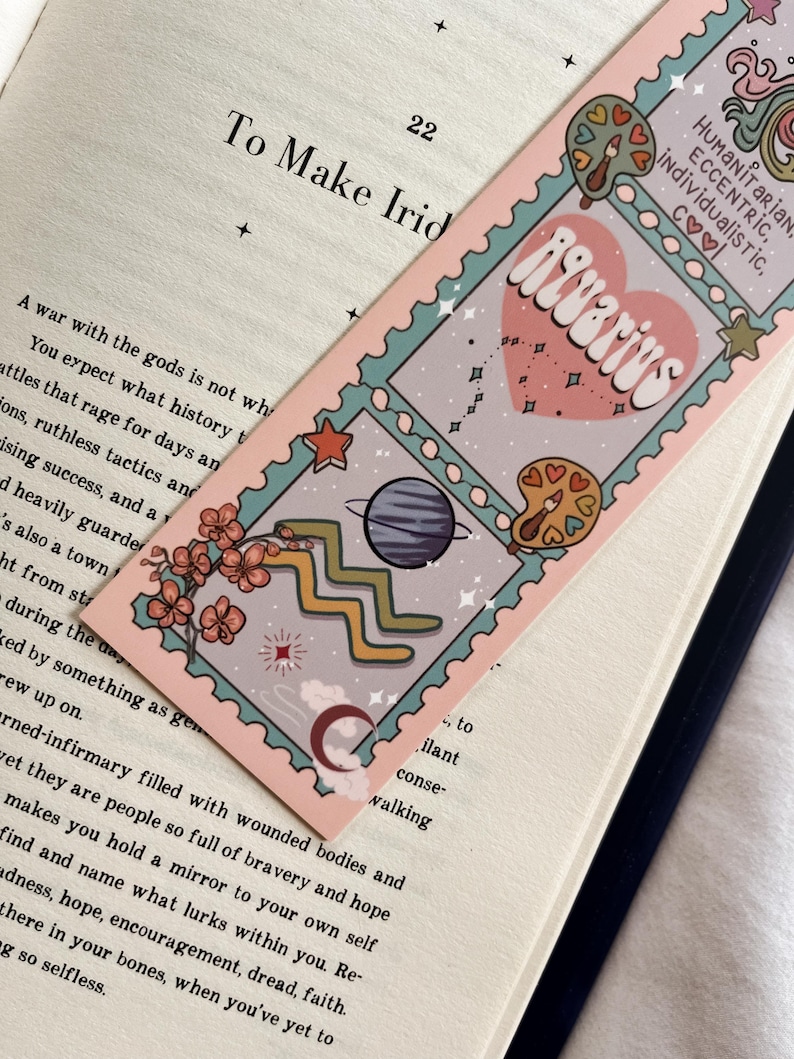 May include: A bookmark with an Aquarius design. The bookmark has a pink border and a light blue interior. The design includes a heart with the word "Aquarius", stars, planets, and other astrological symbols. The bookmark is placed on top of an open book.