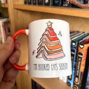 May include: White ceramic mug with an orange handle. The mug features a Christmas tree made of stacked books with a star on top. The text "I'm booked this season" is printed on the mug.