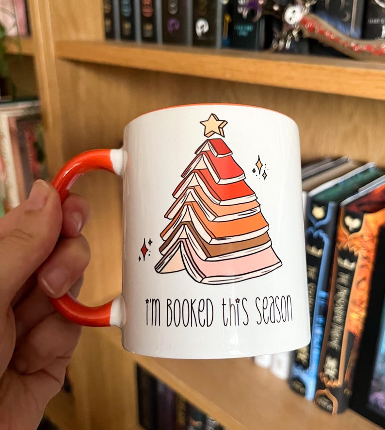 May include: White ceramic mug with an orange handle. The mug features a Christmas tree made of stacked books with a star on top. The text "I'm booked this season" is printed on the mug.
