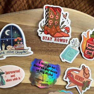K&ouml;nnte beinhalten: Ein Set aus zehn bunten Aufklebern mit verschiedenen Designs, darunter ein Fenster mit einer Katze, die ein Buch liest, ein Paar Cowboystiefel, eine scharfe Sauce, ein UFO, ein Buch mit dem Text "Fairy Smut" und ein Stern mit dem Text "Howdy".