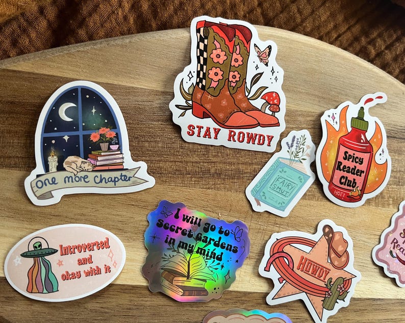 K&ouml;nnte beinhalten: Ein Set aus zehn bunten Aufklebern mit verschiedenen Designs, darunter ein Fenster mit einer Katze, die ein Buch liest, ein Paar Cowboystiefel, eine scharfe Sauce, ein UFO, ein Buch mit dem Text "Fairy Smut" und ein Stern mit dem Text "Howdy".