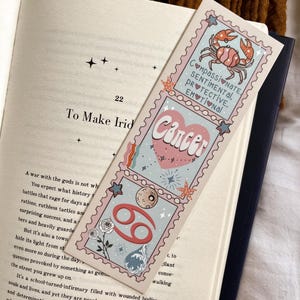 Może przedstawiać: Zakładka w kolorze r&oacute;żowym i niebieskim z ilustracją kraba i tekstem "Compassionate, Sentimental, Protective, Emotional". Zakładka zawiera r&oacute;wnież symbol zodiaku Raka i słowo "Rak" w r&oacute;żowo-białym kolorze.