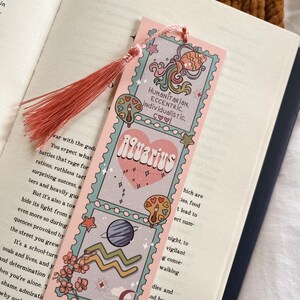 May include: A pink bookmark with an Aquarius theme, featuring illustrations of planets, stars, and the zodiac symbol. The bookmark has a tassel and text that reads "Humanitarian, Eccentric, Individualistic."