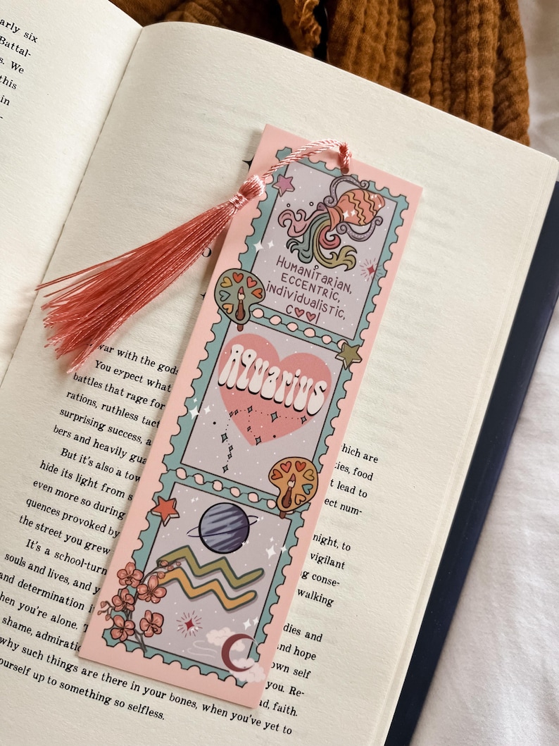 May include: A pink bookmark with an Aquarius theme, featuring illustrations of planets, stars, and the zodiac symbol. The bookmark has a tassel and text that reads "Humanitarian, Eccentric, Individualistic."