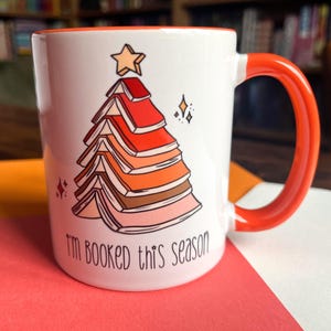 May include: White ceramic mug with an orange handle. The mug features a Christmas tree made of stacked books with a star on top. The text "I'm booked this season" is printed on the mug.