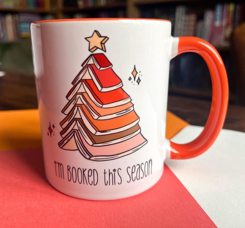 May include: White ceramic mug with an orange handle. The mug features a Christmas tree made of stacked books with a star on top. The text "I'm booked this season" is printed on the mug.