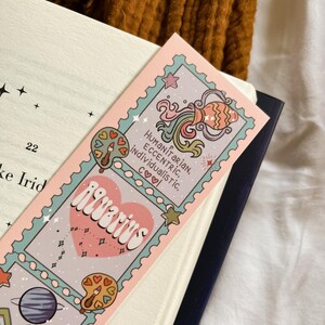 May include: A pink and blue Aquarius zodiac sign bookmark with the words "Aquarius", "Humanitarian, Eccentric, Individualistic, Cool" and illustrations of a planet, a water jug, and a palette. The bookmark is placed in a book.