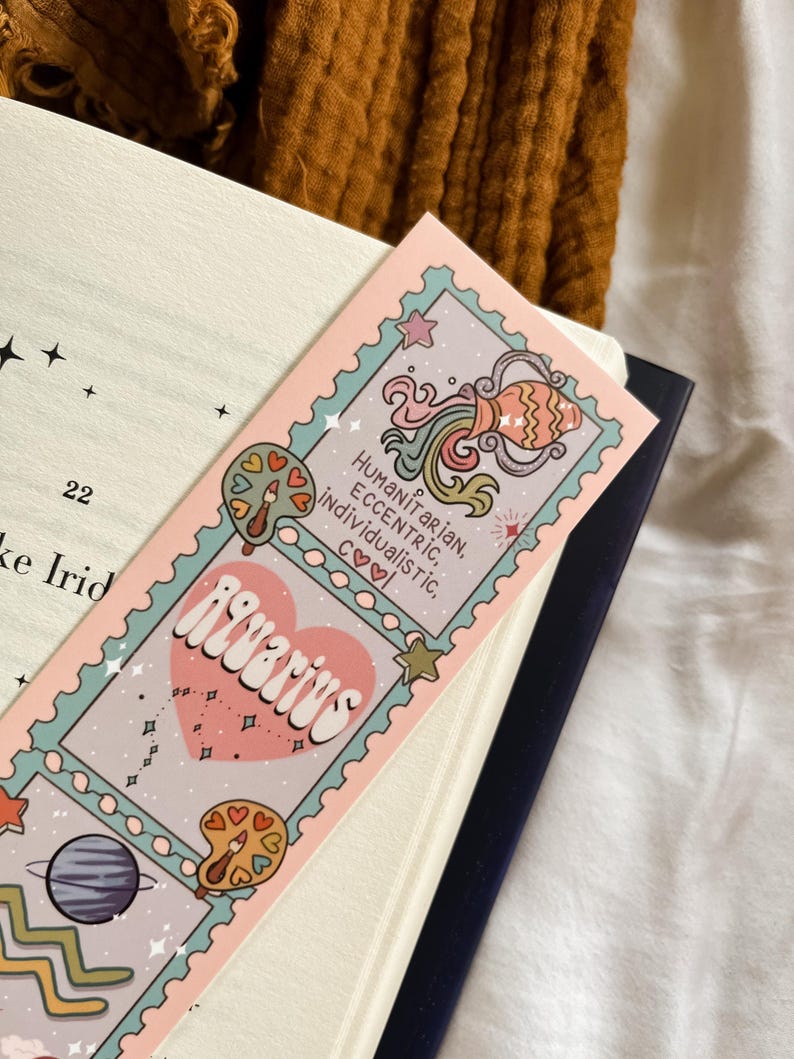 May include: A pink and blue Aquarius zodiac sign bookmark with the words "Aquarius", "Humanitarian, Eccentric, Individualistic, Cool" and illustrations of a planet, a water jug, and a palette. The bookmark is placed in a book.