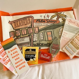 Op de afbeelding: Een cadeaudoos gevuld met boekenthema-artikelen, waaronder een draagtas met een retro motel-illustratie, bladwijzers, een theezakje, een boekrecensiekaart en een sleutelhanger. De draagtas heeft de tekst "Readers Motel" en "Palm Springs".