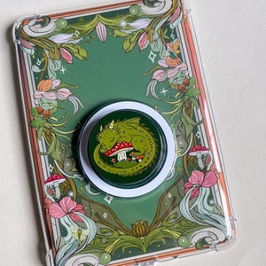 Op de afbeelding: Transparante telefoonhoes met een groen en bloemenontwerp. Een groene draak rust op een rode paddenstoel op de telefoonhouder. Een kleine paddenstoelbedel hangt aan de onderkant. De hoes heeft een groene achtergrond.