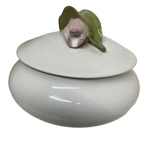 May include: A white ceramic trinket box with a lid, topped with a decorative flower. The flower has a pink center and green leaves. The box is oval-shaped and has a glossy finish.