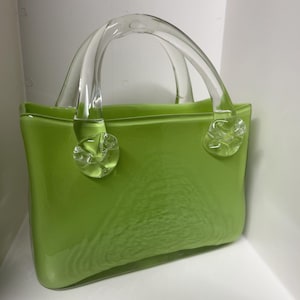 May include: A lime green glass handbag with clear glass handles and decorative floral accents. The bag has a rectangular shape with rounded corners and a glossy finish. The handles are arched and attached to the bag with clear, flower-shaped glass pieces.